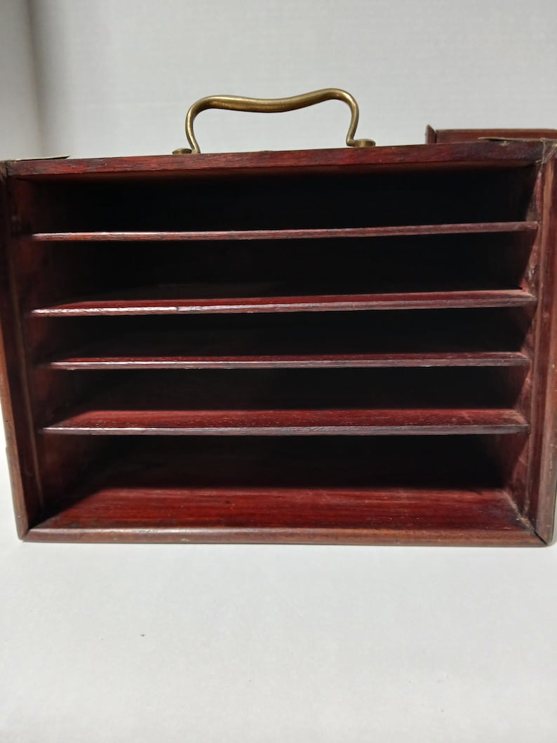 Puede incluir: Un organizador de madera de color marr&oacute;n rojizo oscuro con m&uacute;ltiples ranuras horizontales. Cuenta con un asa de color lat&oacute;n en la parte superior. El organizador parece estar dise&ntilde;ado para guardar documentos o art&iacute;culos peque&ntilde;os, ofreciendo un dise&ntilde;o cl&aacute;sico y funcional.