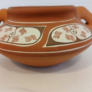 May include: A terracotta-coloured ceramic pot with two loop handles. The pot features a decorative band with cream and brown geometric patterns. The design includes stylised leaf shapes and repeating rectangular motifs. The pot sits on a white surface.