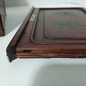 Puede incluir: Primer plano de una bandeja de madera con un acabado marr&oacute;n rojizo oscuro. La bandeja tiene un borde elevado y un centro empotrado con dise&ntilde;os florales pintados de verde. Una parte de una caja de madera es visible a la izquierda.