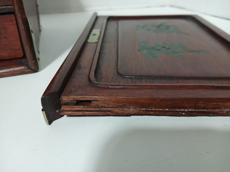 Puede incluir: Primer plano de una bandeja de madera con un acabado marr&oacute;n rojizo oscuro. La bandeja tiene un borde elevado y un centro empotrado con dise&ntilde;os florales pintados de verde. Una parte de una caja de madera es visible a la izquierda.