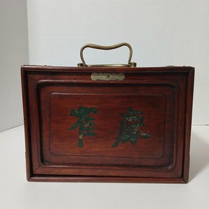 Puede incluir: Una caja rectangular de madera con un asa de lat&oacute;n y letras verdes decorativas. La caja es de color marr&oacute;n oscuro, con un asa de lat&oacute;n y una peque&ntilde;a placa de lat&oacute;n. La rotulaci&oacute;n parece ser caracteres chinos.