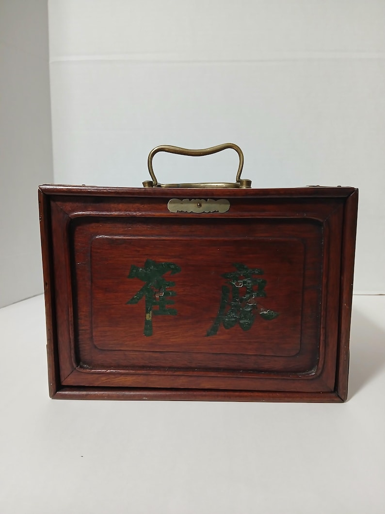 Puede incluir: Una caja rectangular de madera con un asa de lat&oacute;n y letras verdes decorativas. La caja es de color marr&oacute;n oscuro, con un asa de lat&oacute;n y una peque&ntilde;a placa de lat&oacute;n. La rotulaci&oacute;n parece ser caracteres chinos.