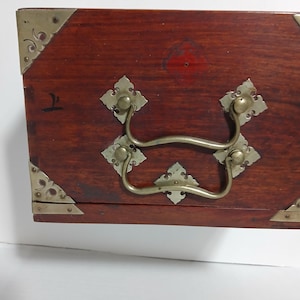 Puede incluir: Una caja rectangular de madera con esquinas de lat&oacute;n y un asa de lat&oacute;n. La caja est&aacute; te&ntilde;ida de un color marr&oacute;n rojizo oscuro. Las esquinas est&aacute;n reforzadas con placas decorativas de lat&oacute;n.