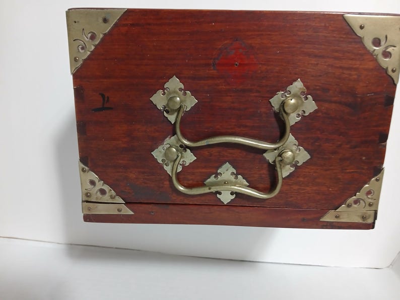 Puede incluir: Una caja rectangular de madera con esquinas de lat&oacute;n y un asa de lat&oacute;n. La caja est&aacute; te&ntilde;ida de un color marr&oacute;n rojizo oscuro. Las esquinas est&aacute;n reforzadas con placas decorativas de lat&oacute;n.