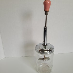 Puede incluir: Un barril de mantequilla vintage de vidrio y cromo con un mango de madera rosa. El barril tiene marcas para 1/3 de taza y 1 taza.