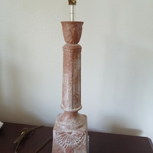 May include: A terracotta-colored table lamp with a column-shaped base and a decorative floral design. The lamp has a square base and a cylindrical column, topped with a bulb socket and a clear glass tube. The lamp is approximately 24 inches tall.