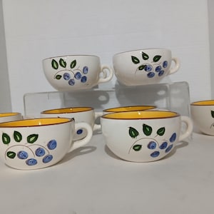 May include: A set of eight white ceramic soup bowls with yellow rims and handles. Each bowl is decorated with a hand-painted design of blueberries and green leaves. The bowls are arranged on a white surface.