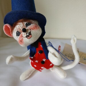 Peut inclure: Une figurine de souris en feutre fantaisiste, coiff&eacute;e d'un haut-de-forme bleu, d'un gilet rouge orn&eacute; d'&eacute;toiles blanches et d'une veste bleue. La souris a un visage souriant avec des joues roses et est pos&eacute;e de mani&egrave;re ludique. Un objet de collection charmant.