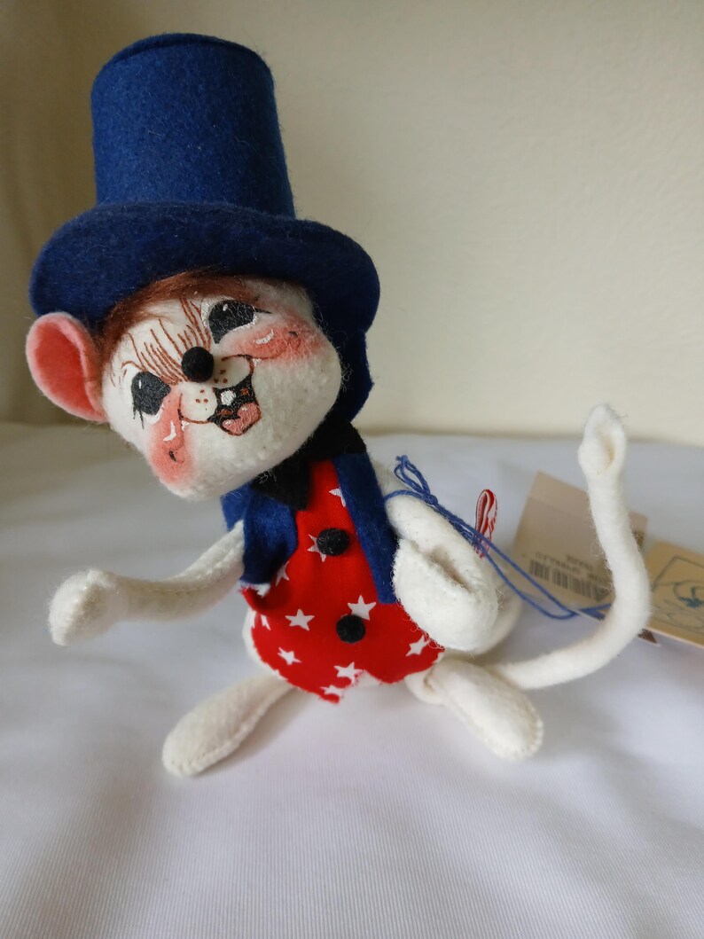 Peut inclure: Une figurine de souris en feutre fantaisiste, coiff&eacute;e d'un haut-de-forme bleu, d'un gilet rouge orn&eacute; d'&eacute;toiles blanches et d'une veste bleue. La souris a un visage souriant avec des joues roses et est pos&eacute;e de mani&egrave;re ludique. Un objet de collection charmant.