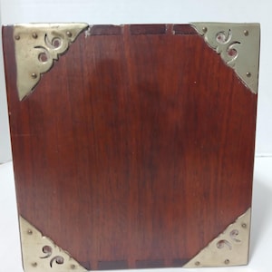 Puede incluir: Una caja de madera cuadrada de color marr&oacute;n oscuro. Las esquinas est&aacute;n adornadas con protectores de esquina decorativos de metal dorado. La caja tiene un acabado pulido y parece estar hecha de madera maciza.