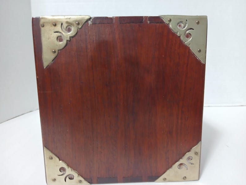 Puede incluir: Una caja de madera cuadrada de color marr&oacute;n oscuro. Las esquinas est&aacute;n adornadas con protectores de esquina decorativos de metal dorado. La caja tiene un acabado pulido y parece estar hecha de madera maciza.