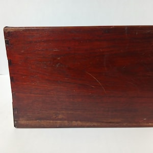 Puede incluir: Una caja rectangular de madera con un acabado marr&oacute;n rojizo intenso. La caja est&aacute; construida con juntas de cola de milano visibles en las esquinas, lo que indica un dise&ntilde;o artesanal. La veta de la madera es prominente y la caja parece envejecida.