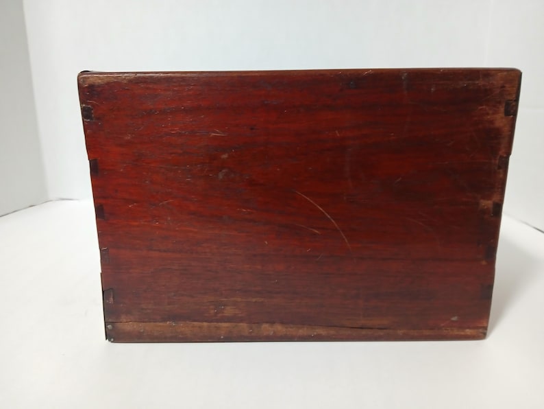 Puede incluir: Una caja rectangular de madera con un acabado marr&oacute;n rojizo intenso. La caja est&aacute; construida con juntas de cola de milano visibles en las esquinas, lo que indica un dise&ntilde;o artesanal. La veta de la madera es prominente y la caja parece envejecida.