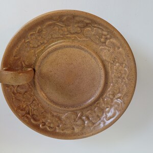 Può includere: Un piattino in ceramica marrone con un motivo floreale in rilievo attorno al bordo e un piccolo manico sul lato.