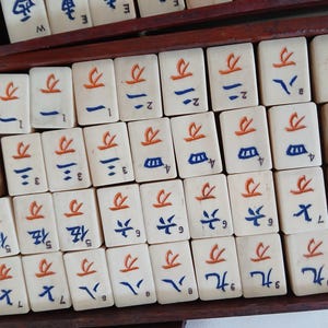Puede incluir: Un conjunto de fichas de Mahjong de color marfil dispuestas en una caja de madera. Cada ficha presenta caracteres y s&iacute;mbolos chinos en rojo y azul. Las fichas rectangulares tienen una superficie lisa, con varios n&uacute;meros y s&iacute;mbolos.