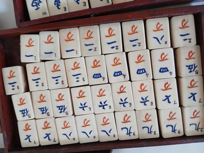 Puede incluir: Un conjunto de fichas de Mahjong de color marfil dispuestas en una caja de madera. Cada ficha presenta caracteres y s&iacute;mbolos chinos en rojo y azul. Las fichas rectangulares tienen una superficie lisa, con varios n&uacute;meros y s&iacute;mbolos.