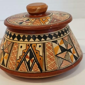 May include: A hand-painted ceramic jar with a lid. The jar is brown with geometric patterns in black, orange, and cream. The lid has a small knob and similar decorative elements. The jar is likely used for storage.