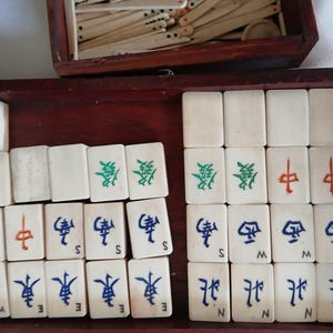 Puede incluir: Un juego de Mahjong vintage con fichas de color marfil dispuestas en una caja de madera. Las fichas presentan caracteres chinos en verde, rojo y azul. Un compartimento separado contiene palos de madera adicionales. La caja es de color marr&oacute;n rojizo oscuro.