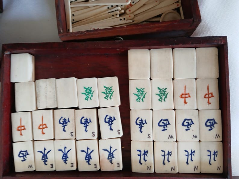 Puede incluir: Un juego de Mahjong vintage con fichas de color marfil dispuestas en una caja de madera. Las fichas presentan caracteres chinos en verde, rojo y azul. Un compartimento separado contiene palos de madera adicionales. La caja es de color marr&oacute;n rojizo oscuro.