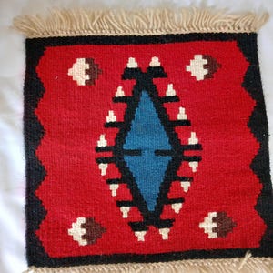 May include: A small, square woven rug with a vibrant red background. The rug features a central blue diamond shape, surrounded by white and brown geometric patterns. Black borders and beige fringe complete the design, showcasing a traditional, handcrafted aesthetic.