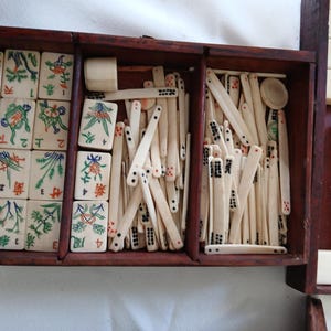 Puede incluir: Un juego de Mahjong vintage en una caja de madera marr&oacute;n oscuro. El juego incluye fichas de color marfil con ilustraciones coloridas y caracteres chinos, adem&aacute;s de palos de madera. La caja tiene varios compartimentos.