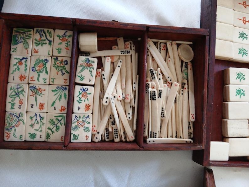 Puede incluir: Un juego de Mahjong vintage en una caja de madera marr&oacute;n oscuro. El juego incluye fichas de color marfil con ilustraciones coloridas y caracteres chinos, adem&aacute;s de palos de madera. La caja tiene varios compartimentos.