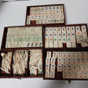 Puede incluir: Un juego de Mahjong vintage con fichas de color marfil y cajas de almacenamiento de madera. Las fichas presentan caracteres chinos y s&iacute;mbolos coloridos. El juego incluye varias fichas y palos.