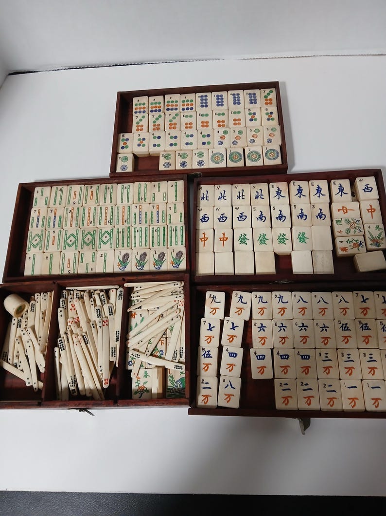 Puede incluir: Un juego de Mahjong vintage con fichas de color marfil y cajas de almacenamiento de madera. Las fichas presentan caracteres chinos y s&iacute;mbolos coloridos. El juego incluye varias fichas y palos.