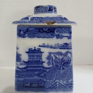 May include: A blue and white ceramic tea caddy with a pagoda design. The rectangular caddy has a matching lid with a detailed pattern. The caddy features a landscape scene with trees and buildings.