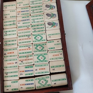 Puede incluir: Un juego de Mahjong vintage con fichas de color marfil dispuestas en una bandeja de madera. Las fichas presentan caracteres chinos, n&uacute;meros y s&iacute;mbolos coloridos en verde, rojo y azul. El juego incluye una variedad de fichas.