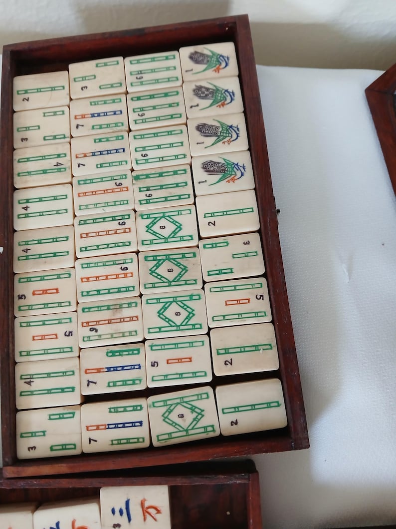 Puede incluir: Un juego de Mahjong vintage con fichas de color marfil dispuestas en una bandeja de madera. Las fichas presentan caracteres chinos, n&uacute;meros y s&iacute;mbolos coloridos en verde, rojo y azul. El juego incluye una variedad de fichas.