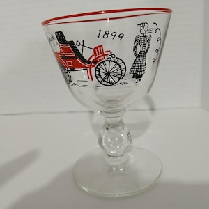 Puede incluir: Una copa de vidrio transparente con un borde rojo y una ilustración en blanco y negro de un carruaje tirado por caballos y una mujer con un vestido. La ilustración incluye el año "1899".