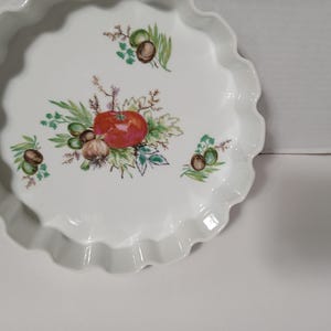 May include: White ceramic pie dish with a scalloped edge. The center features a colorful illustration of a red tomato, olives, garlic, and herbs. The dish is designed for baking and serving.