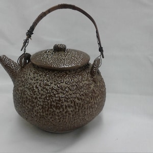 Può includere: Una teiera in ceramica marrone con superficie strutturata e manico intrecciato. Il coperchio ha un motivo strutturato abbinato e un piccolo pomello. Il beccuccio è sul lato sinistro. Il manico è in materiale marrone scuro.