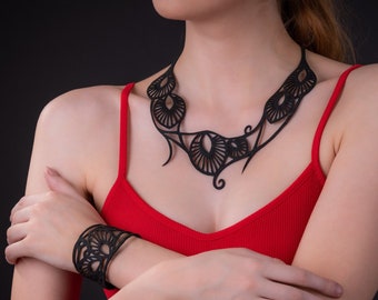May include: A black, laser-cut necklace and matching cuff bracelet. The jewelry features an openwork design with floral and geometric shapes. The woman in the image is wearing a red tank top against a black background.