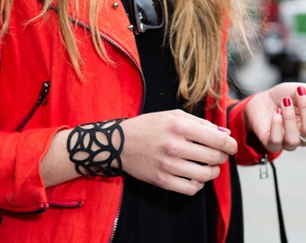 May include: A woman wearing a red jacket with a black leather bracelet with a geometric design. The bracelet is on her left wrist.