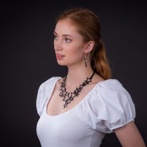 May include: A woman with red hair wearing a white blouse with a black star-shaped necklace and earrings.