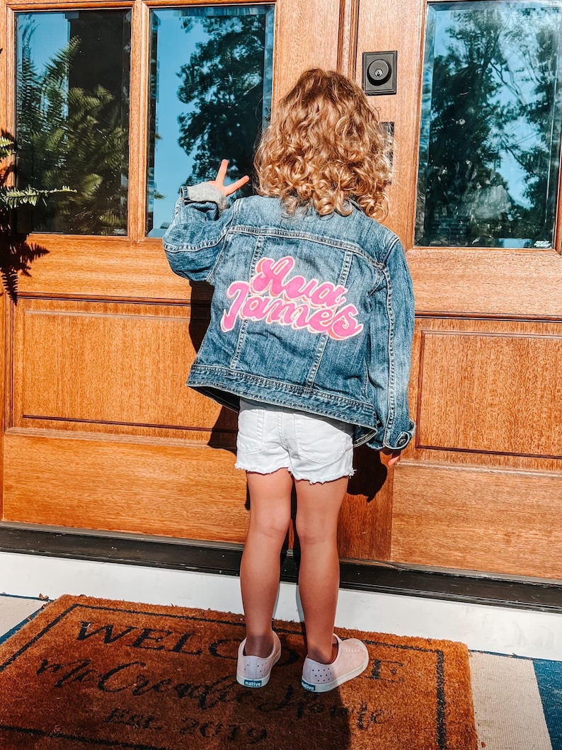 May include: A young person wearing a blue denim jacket with a pink "Ava James" patch on the back. They are standing in front of a wooden door with a welcome mat.