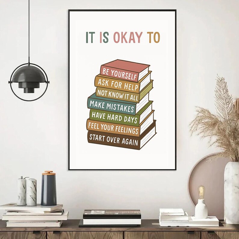 May include: A framed print with the text "IT IS OKAY TO" above a stack of illustrated books. Each book has a different color and title, including "BE YOURSELF" and "START OVER AGAIN". The print is in a black frame.
