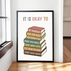 May include: A framed print with the words "IT IS OKAY TO" at the top, above a stack of illustrated books. Each book has a different title, including "BE YOURSELF" and "START OVER AGAIN". The frame is black.