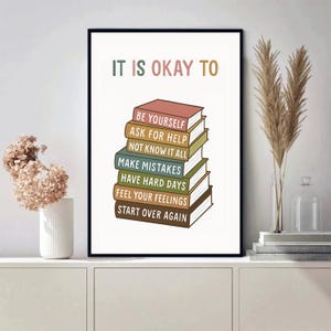 May include: A framed art print with the text "IT IS OKAY TO" above a stack of colorful books. Each book spine has a different phrase, including "BE YOURSELF" and "START OVER AGAIN". The artwork is displayed on a white surface.