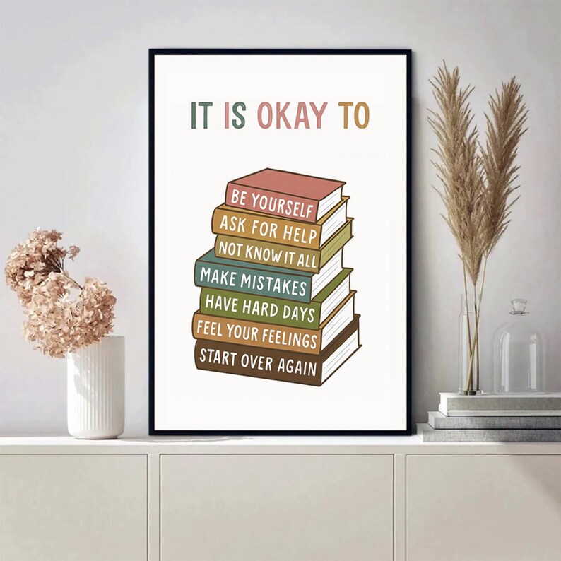 May include: A framed art print with the text "IT IS OKAY TO" above a stack of colorful books. Each book spine has a different phrase, including "BE YOURSELF" and "START OVER AGAIN". The artwork is displayed on a white surface.