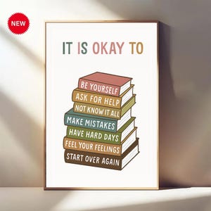 May include: A framed print with the text "IT IS OKAY TO" above a stack of illustrated books. Each book spine has a different phrase, including "BE YOURSELF" and "START OVER AGAIN." A red "NEW" sticker is in the upper left corner.