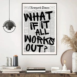 May include: A framed print featuring a newspaper-style background with the headline "WHAT IF IT ALL WORKS OUT?" in bold, black lettering. The print is displayed on a white wall above a wooden cabinet, with decorative items.