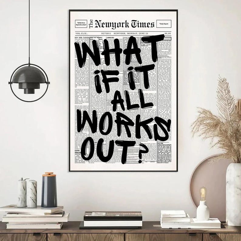 May include: A framed print featuring a newspaper-style background with the headline "WHAT IF IT ALL WORKS OUT?" in bold, black lettering. The print is displayed on a white wall above a wooden cabinet, with decorative items.