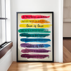 May include: A framed print with a rainbow background and text that reads "In this home, we believe NO HUMAN IS ILLEGAL love is love BLACK LIVES MATTER feminism is for everyone ALL BODIES ARE BEAUTIFUL water is life SCIENCE IS REAL and compassion is everything."