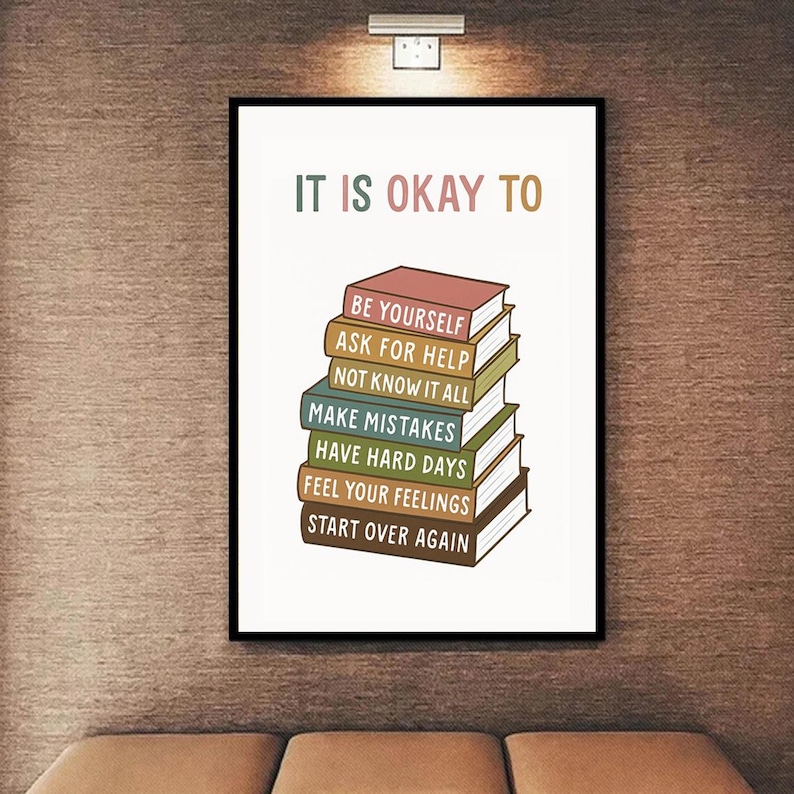 May include: A framed print with the words "IT IS OKAY TO" at the top. Below, a stack of illustrated books with phrases like "BE YOURSELF" and "START OVER AGAIN". The frame is black, and the background is a textured brown.
