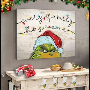 May include: A rectangular canvas print featuring a wood-grain background with the text "every family has one" written in black script. A string of colorful Christmas lights hangs above a green character wearing a red and white Santa hat.