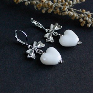 May include: A pair of silver-toned bow earrings with white heart-shaped pendants. The earrings have a lever back closure. The background is a dark, solid color, with a sprig of dried flowers.