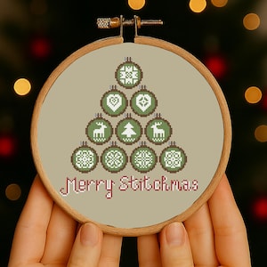 May include: A cross-stitch embroidery hoop featuring a Christmas tree design made of green ornaments with various patterns. The text "Merry Stitchmas" is stitched in pink. The hoop is held in front of a blurred Christmas tree.
